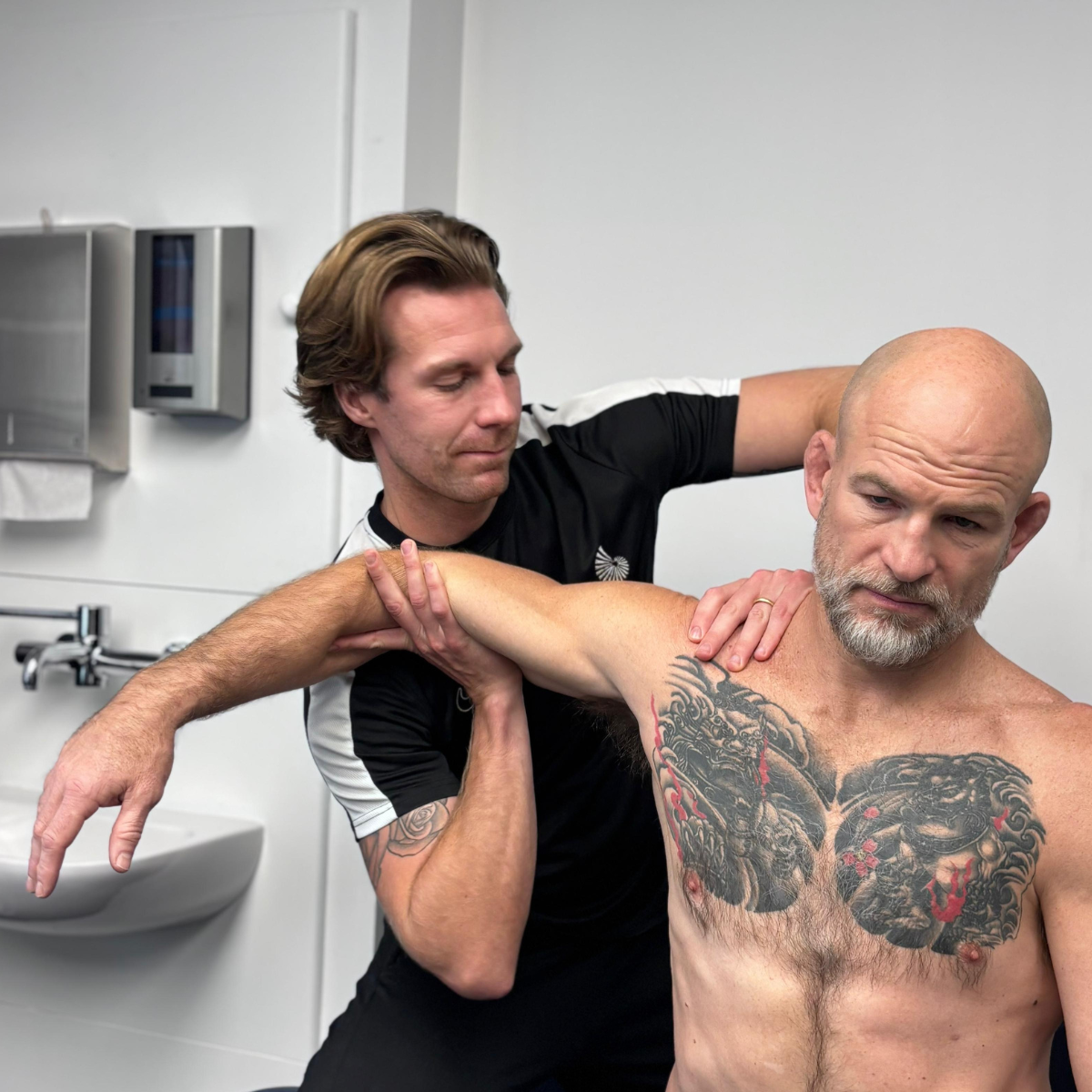 Physiotherapist supporting a shirtless male patient’s arm and shoulder during a mobility assessment in a clinical treatment room, with the patient showing a large chest tattoo.