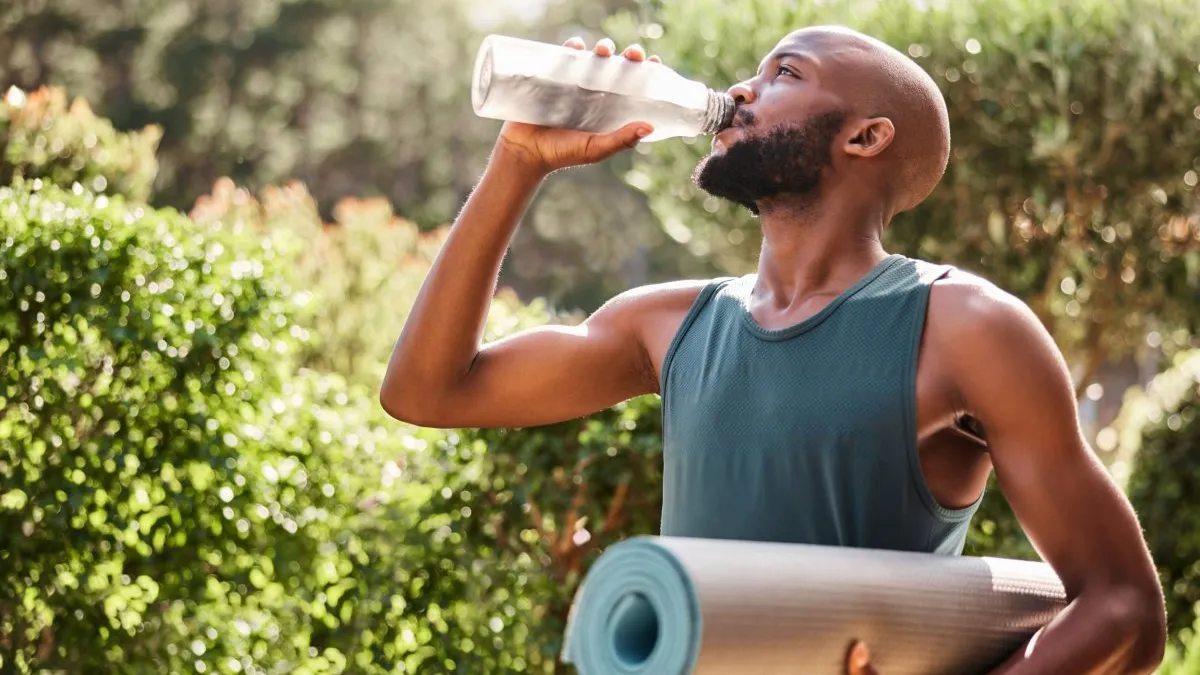 Man drinking water outdoors while carrying an exercise mat